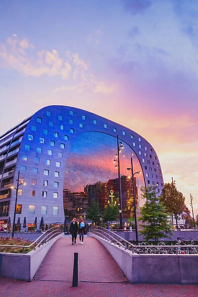 A couple crossing a bridge, at sunset in front of the Market Hall in Rotterdam on a
