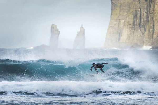 A guy surfing along the coast in front of Tjornuvik. In the background the Risin og Kellingin sea stacks. Streymoy, Faroe Islands