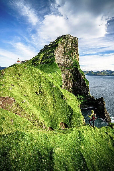 Hiker admiring Kallur lighthouse and Borgarin mountain peak standing on top of cliffs, Kalsoy island, Faroe Islands, Denmark