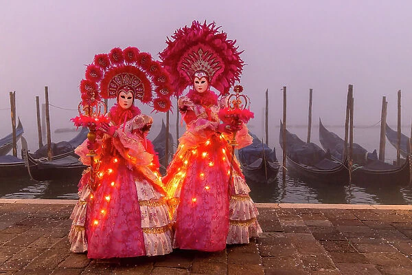 Italy, Veneto, Venice, two models in illuminated costumes pose in front of gondolas on a foggy morning during the Venice Carnival