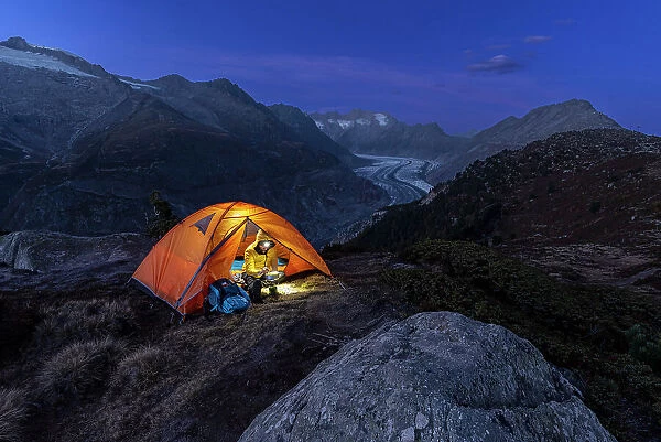 Man with head torch preparing meals outside the tent pitched above the Aletsch glacier at dusk, Bernese Alps, Valais canton, Switzerland (MR)