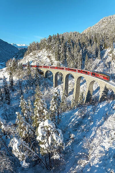 Red train crossing a viaduct in the fairy tale snowy landscape under a clear winter sky, Graubunden canton, Switzerland