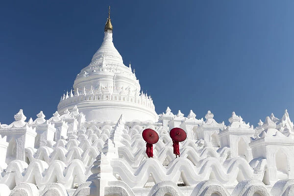 Two young Buddhist monks stand on the white walls of Hsinbyume Pagoda holding red