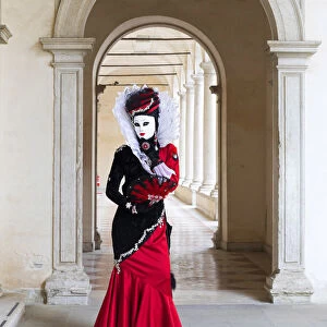 A woman in a red costume and mask poses in an archway during the Venice Carnival