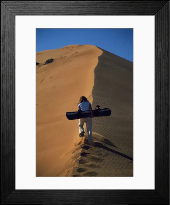 Framed Print : An adventurer climbs a sand dune with a sand board Framed Print : An adventurer climbs a sand dune with a sand board