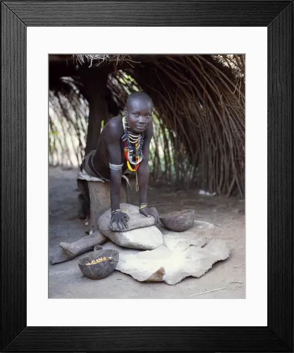 Framed Print : A Kwego woman grinds sorghum flour at the entrance to her hut Framed Print : A Kwego woman grinds sorghum flour at the entrance to her hut