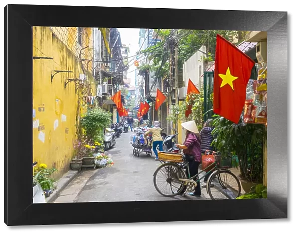 Woman pusing a bicycle through an alleyway lined with red Vietnamese flags, Hoan