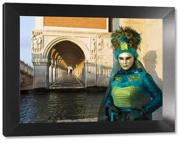 A woman in a mask and green costume stands by the canal next to St Marks square