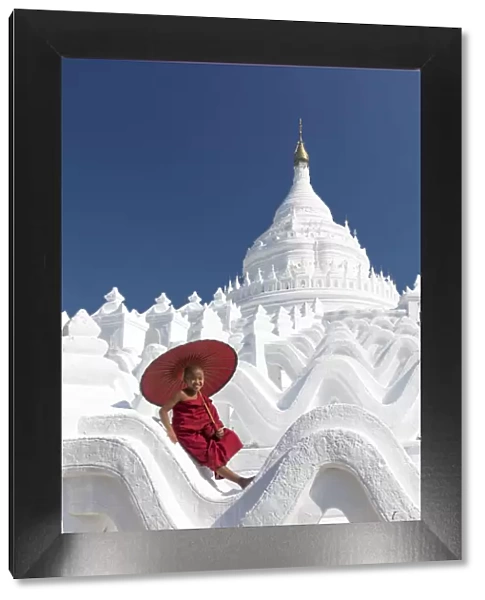 Novice monk sits on the white wall of Hsinbyume Pagoda holding a red umbrella, Mingun