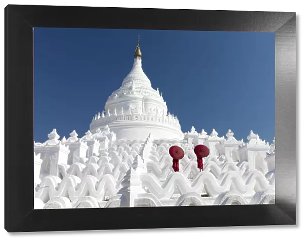 Two young Buddhist monks stand on the white walls of Hsinbyume Pagoda holding red