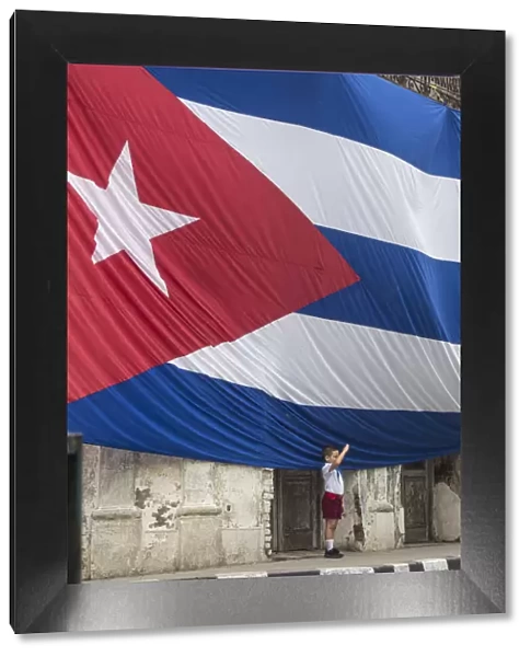 Cuba, Boy saluting to Huge Cuban flag hanging across buildings in a street in Santa Clara
