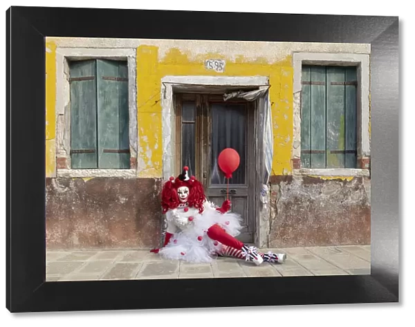 A woman dressed as a clown holds a ballon in front of a colourful facade on Burano