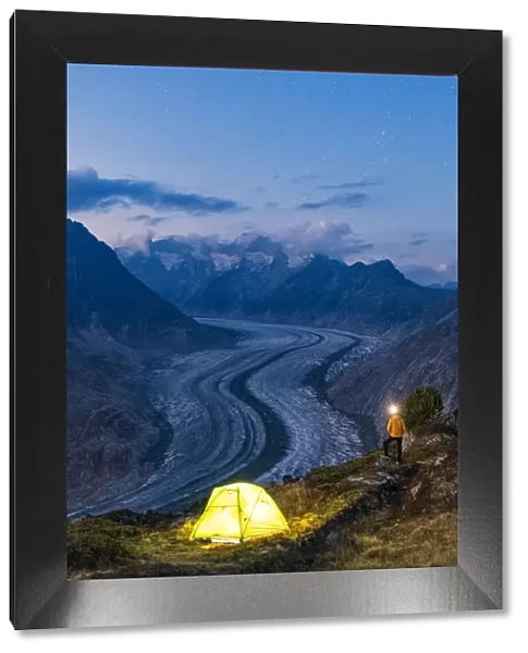 Hiker man with head torch light admiring Aletsch Glacier standing beside the tent