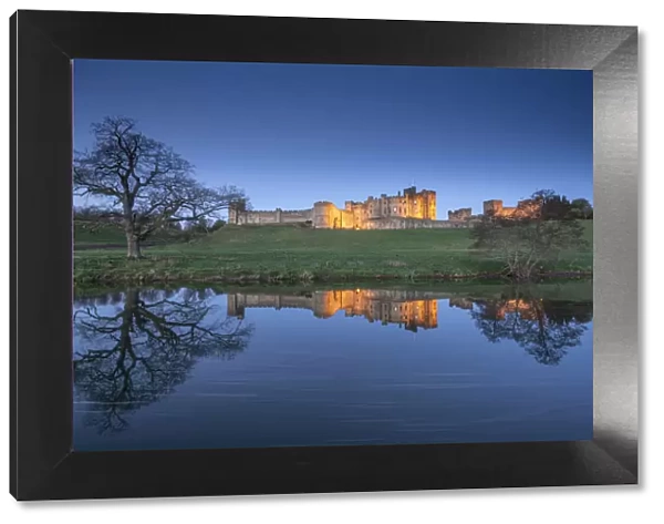 Alnwick Castle illuminated at twilight and reflected in the River Aln, Northumberland