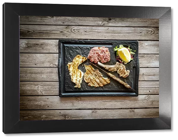 Mixed grilled meat in black tray on rustic wood table background from above, Italy