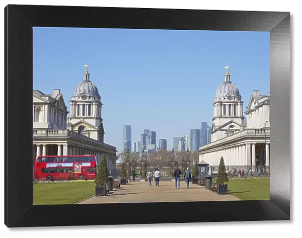 A traditional double-decker red bus passing by the Greenwich University at the Old Royal Naval College with the Canary Wharf skyline in background, London, United Kingdom, Northern Europe