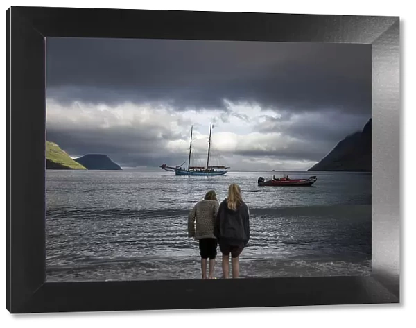 Two girls at G! Festival in Syðrugota walking into the ocean. In the background the sailship Norðlysið. Island of Eysturoy. Faroe Islands