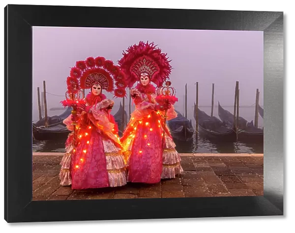 Italy, Veneto, Venice, two models in illuminated costumes pose in front of gondolas on a foggy morning during the Venice Carnival