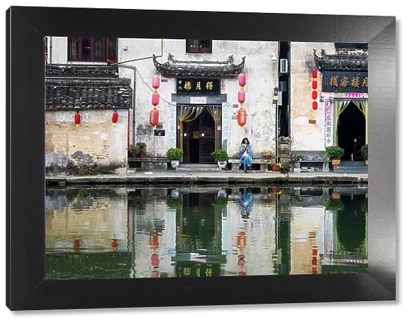 Woman on bench and village buildings reflected in pond, Hongcun, China