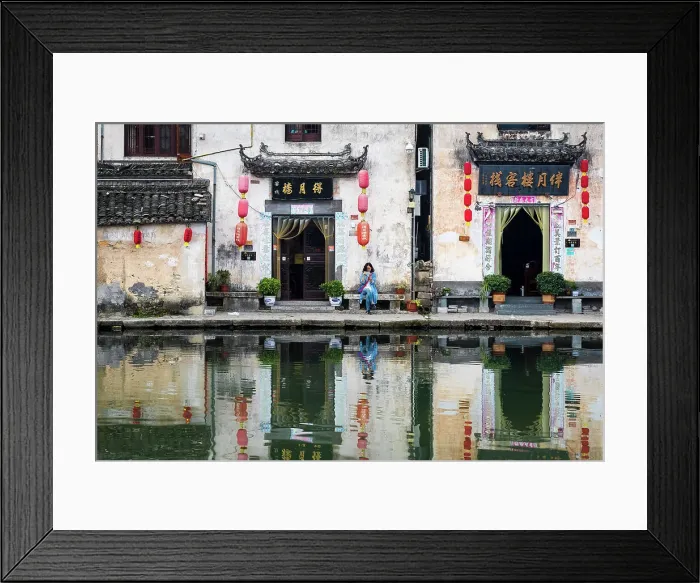 Framed Print : Woman on bench and village buildings reflected in pond, Hongcun, China