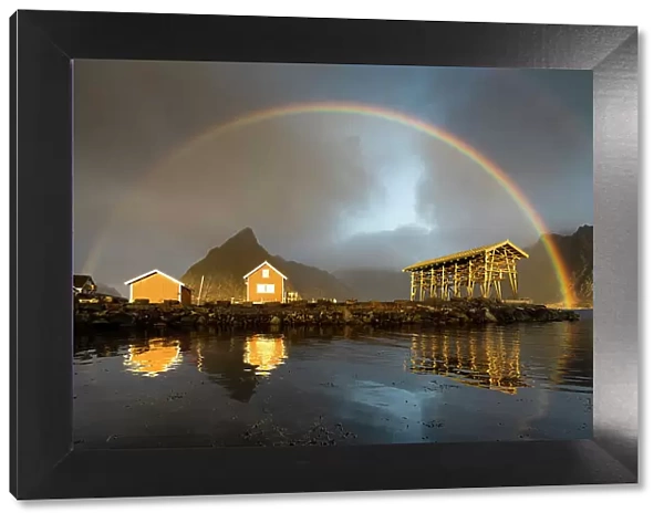 Fishermen cabins under a dramatic cloudy sky with rainbow at dawn, Sakrisoy, Reine, Nordland county, Lofoten Islands, Norway