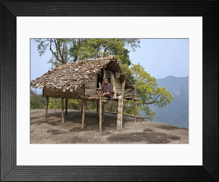 Framed Print : A man sits on the veranda of his small house, raised on stilts, situated precariously on the edge of a Framed Print : A man sits on the veranda of his small house, raised on stilts, situated precariously on the edge of a