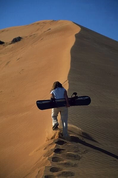 An adventurer climbs a sand dune with a sand board