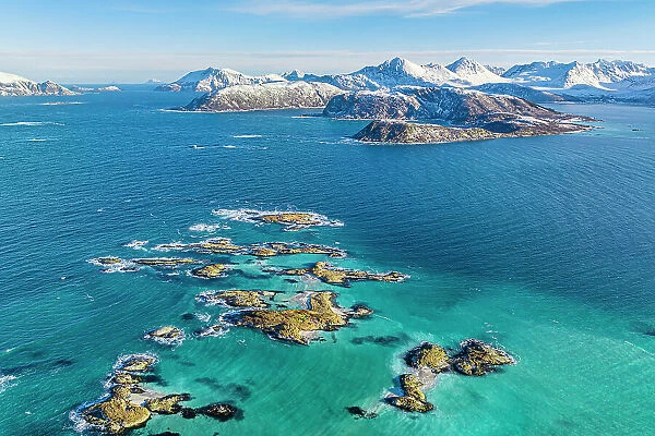 Aerial view of cliffs in the turquoise water of the cold sea with snowy mountains on background, Sommaroy, Troms county, Norway