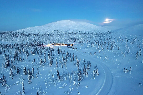 Aerial view of a snowy mountain road in the frozen forest nearby a ski area at dusk, Yllastunturi National Park, Lapland, Finland