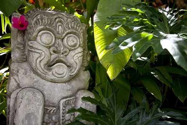 Bali, Ubud. A stone carving, adorned with a hibiscus flower, sits in tropical gardens