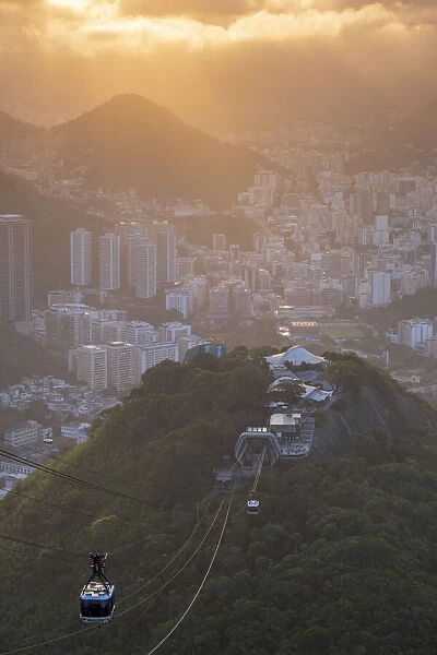Brazil, Rio de Janeiro, Cable cars ascending to Sugar Loaf mountain. Morro da Urca hill, forest, Rio city in the background under golden light