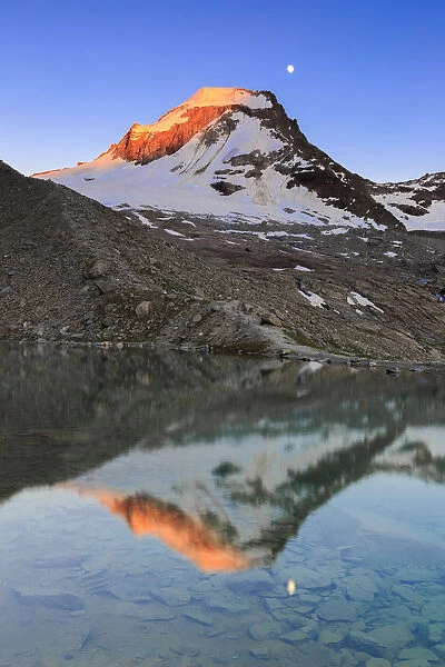 Ciarforon still illuminated by the colors of dawn and the moon are reflected in Moncorv√® lake. Valsavarance, Gran Paradiso National Park, Aosta Valley, Italy