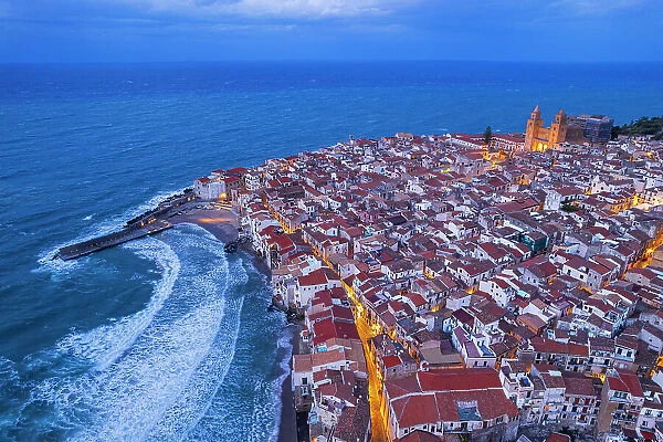 Dusk above the red tile roofs of Cefalu with street lights on and rough sea, aerial view, Cefalu, Palermo province, Tyrrhenian sea, Sicily, Italy