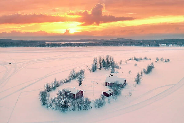 Europe, Finland, Rovaniemi, aerial view of a frozen lake near Rovaniemi with traditional red buildings with the sun rising on a winter morning