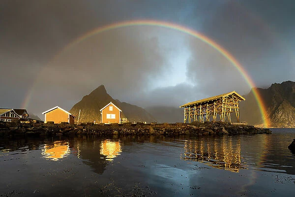 Fishermen cabins under a dramatic cloudy sky with rainbow at dawn, Sakrisoy, Reine, Nordland county, Lofoten Islands, Norway