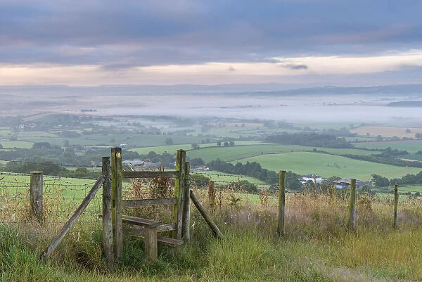 Footpath and stile through farmland with mist covered countryside beyond, Devon, England