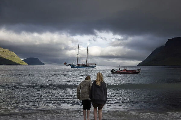 Two girls at G! Festival in Syðrugota walking into the ocean. In the background the sailship Norðlysið. Island of Eysturoy. Faroe Islands