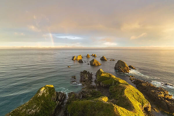 Golden light on The Nuggets islands and rainbow in the sky at Nugget Point. Ahuriri Flat