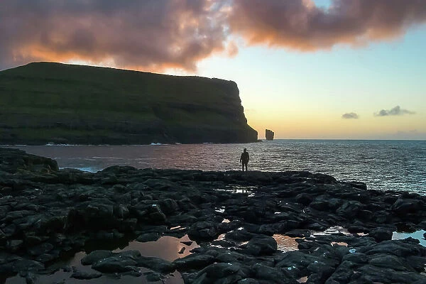 A hiker standing in front of the coast of the village of Eiði at sunset. In the backgroud the Rising and Kellingin sea stacks. Island of Eysturoy. Faroe Islands