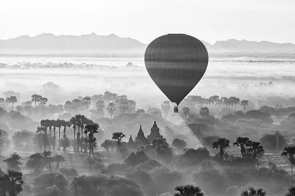 A hot air balloon flies over a trees and a temple at sunrise on a misty morning, Bagan
