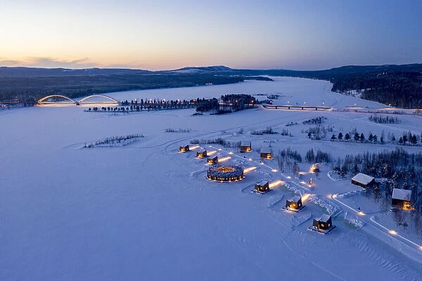 Illuminated floating circular building and wood cabins of Arctic Bath Hotel in the snow at dusk, Harads, Lapland, Sweden