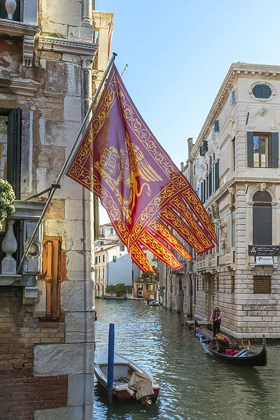 Italy, Veneto, Venice. Gondola passing on a canal with venetian flag hanging from a wall