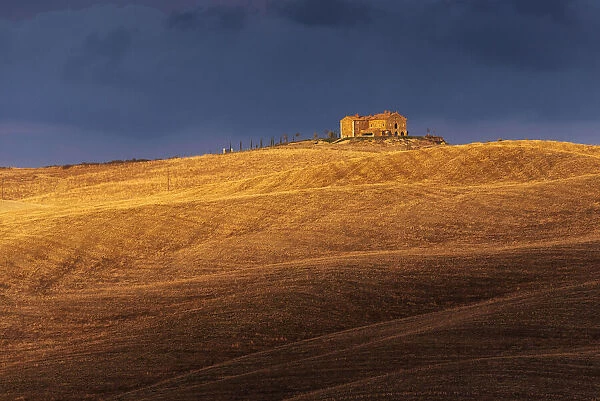 A lonely countryhouse and some rolling hills illuminated by the warm sunset light in autumn, right before a storm rolled in, not far from Montalcino, Val d Orcia, Tuscany, Italy