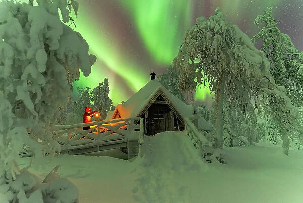 Man with lantern approacching a wooden hut in the snow covered forest under northern lights (aurora borealis), Lapland, Finland (MR)