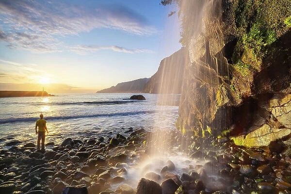 A man observes sunrise at a waterfall in Seixal black sand beach, Seixal, Porto Moniz, Madeira, Portugal (MR)