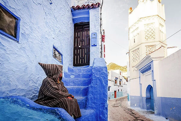 Man with traditional Djellaba looking at white tower of minaret sitting in the blue old town of Chefchaouen, Morocco