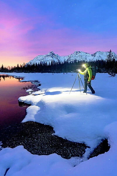 Man with tripod photographing lake Entova at dawn standing on the snowy shores, Valmalenco, Valtellina, Lombardy, Italy