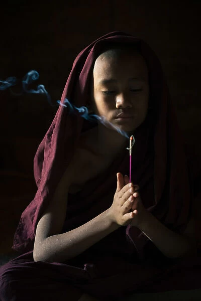 Novice monk holding a lit incense stick while praying inside a temple, UNESCO, Bagan