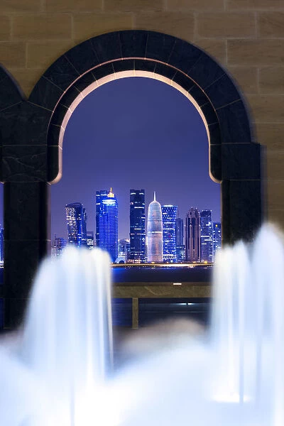 Qatar, Doha. Cityscape at night framed in a arch with fountain in the foreground