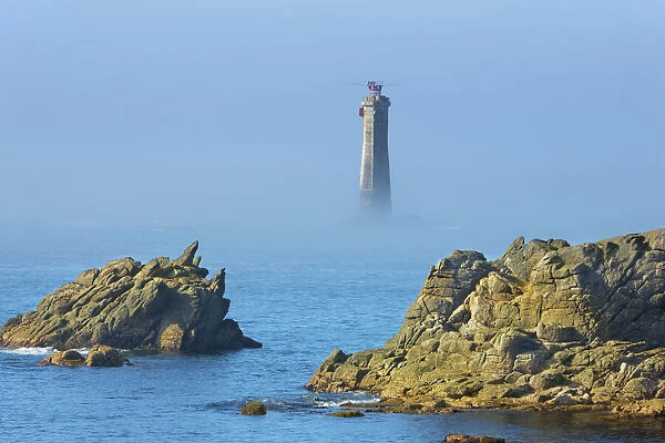 Rocky coast at Pointe de Pern with lighthouse of Nividic in fog - France, Brittany